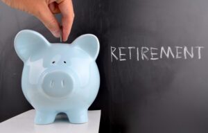 A hand putting a coin into a blue piggybank. A blackboard says "retirement" in chalk writing behind it. Average 60-year Old Have Saved for Retirement?