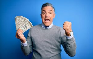 A man in a gray sweatshirt holds up a fan of cash as he owns the best performing stocks.